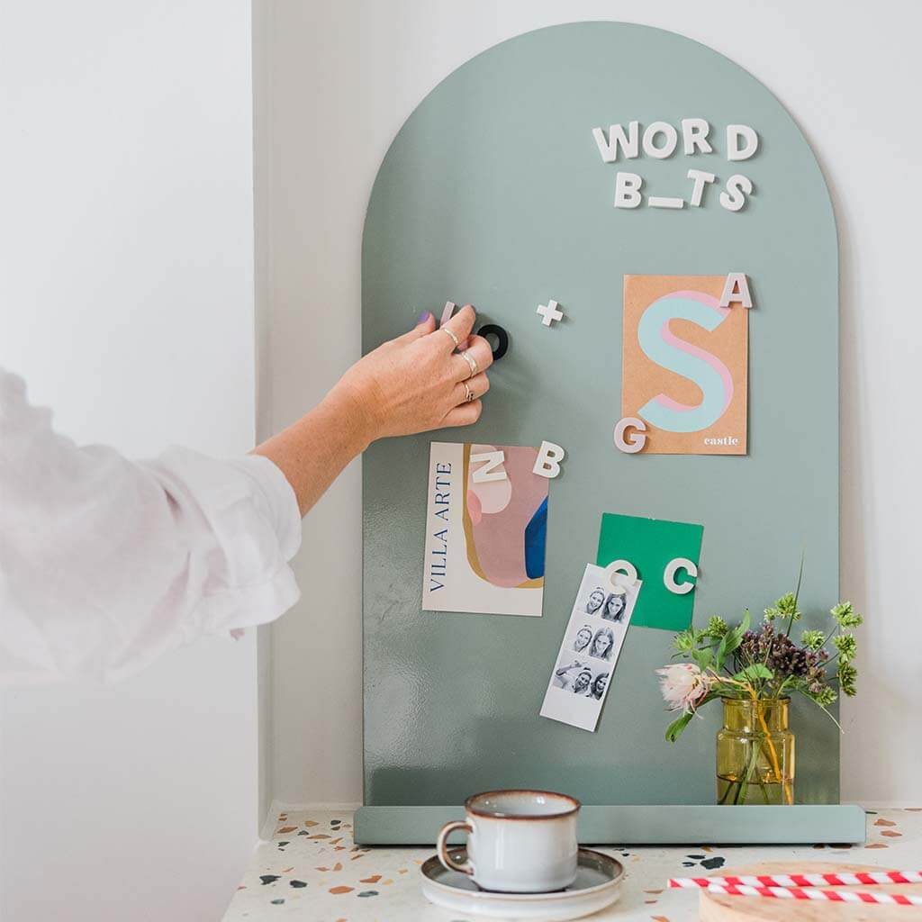 Sage green Moodyboard magnet board arch with bone, black and blush Wordbits magnetic letters on it and the hand of a woman moving a letter